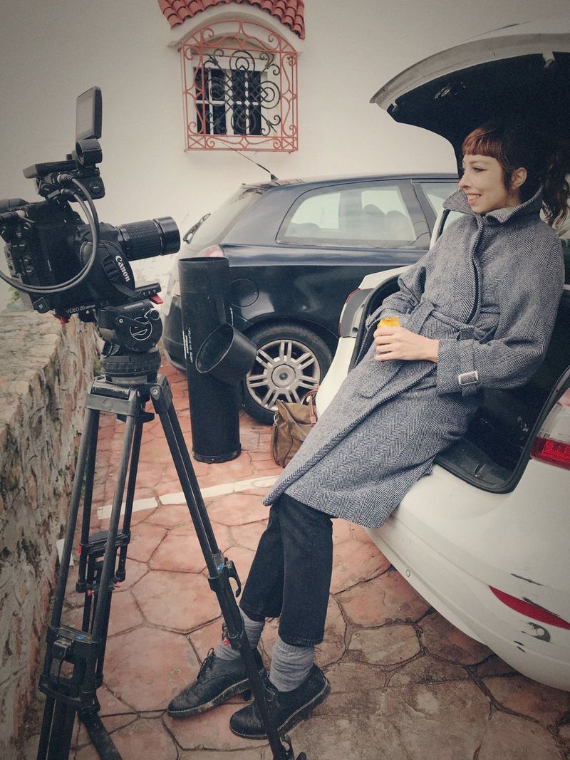 Filmmaker Irene Cruz smiles as she leans against the open boot of a car. A Canon camera is set up on a tripod in front of her.