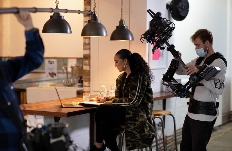 Director Ashleigh Jadee sits at a breakfast bar while a camera films her. She is looking at her laptop and smiling, while writing in her notepad.
