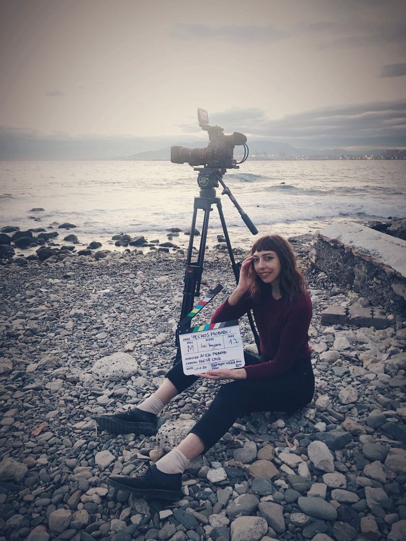 Cinematographer Irene Cruz sits on a rocky beach next to a Canon camera on a tripod. She is holding a clapperboard.