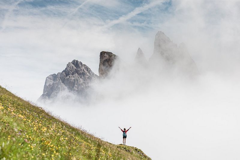 A woman with her arms raised in the air, standing on a steep, grassy hill, with rocky crags rising into view through the clouds in the background.