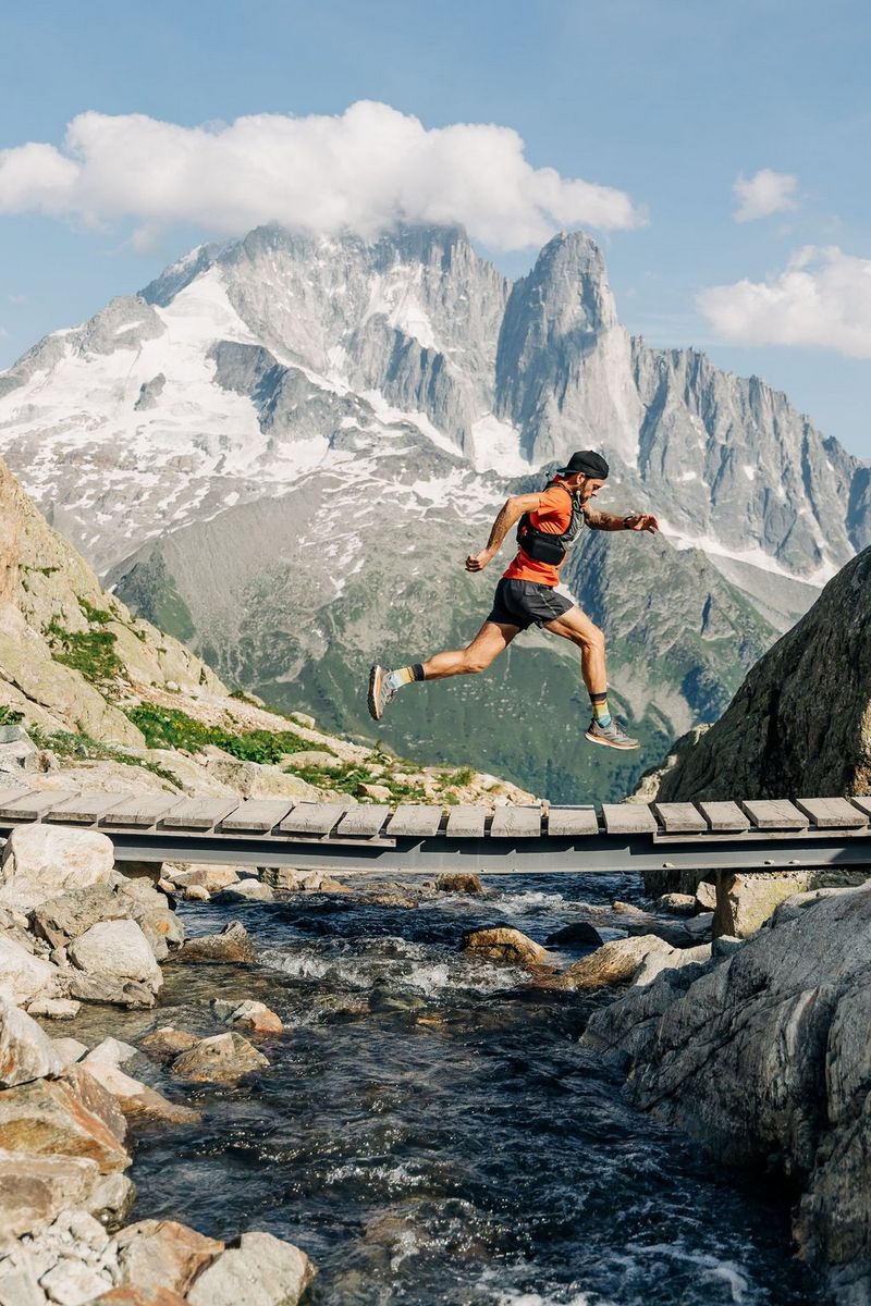 A man captured in mid air as he runs over a wooden bridge over a stream in front of Mont Blanc.