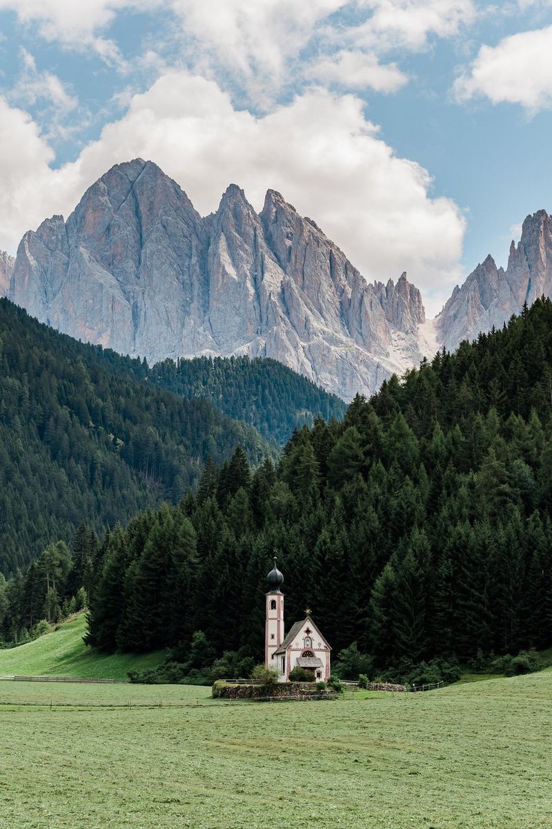 A church with a tower topped with an onion dome sits isolated on the edge of a dense forest with steep mountains in the background.