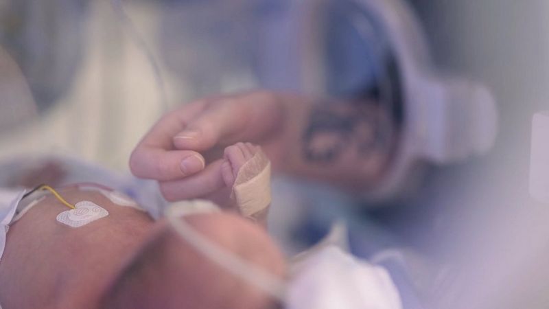 In a still from a video shot on a Canon EOS C70, an adult hand rests inside a baby incubator, the tiny hand of the baby holding on to one finger. 