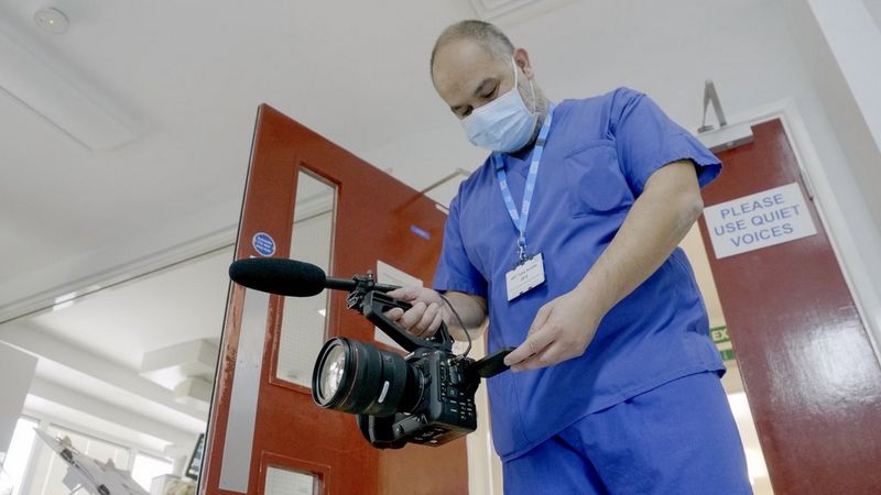 Filmmaker Giulio Di Sturco stands in blue hospital scrubs, looking down at the screen of a Canon EOS C70 camera that he is holding at waist level in front of him.