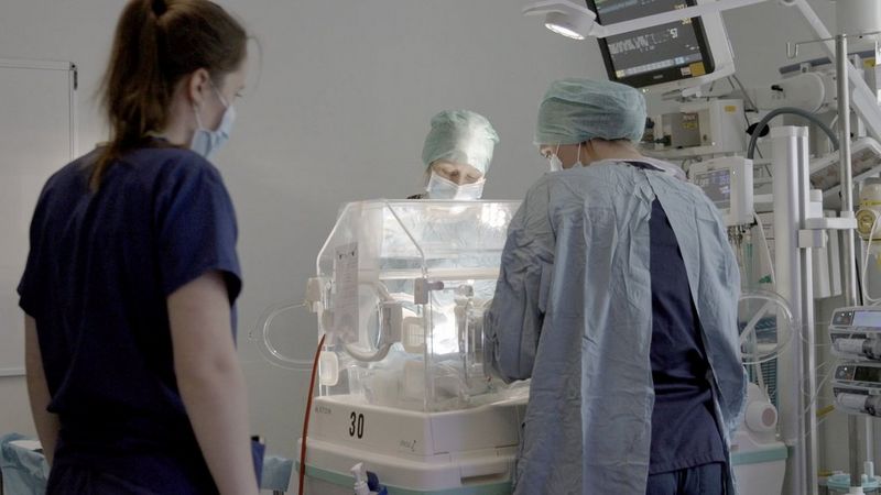 A still from a video shot on a Canon EOS C70, showing three healthcare workers standing around a baby incubator looking in, with lots of hospital equipment and monitors at one side.