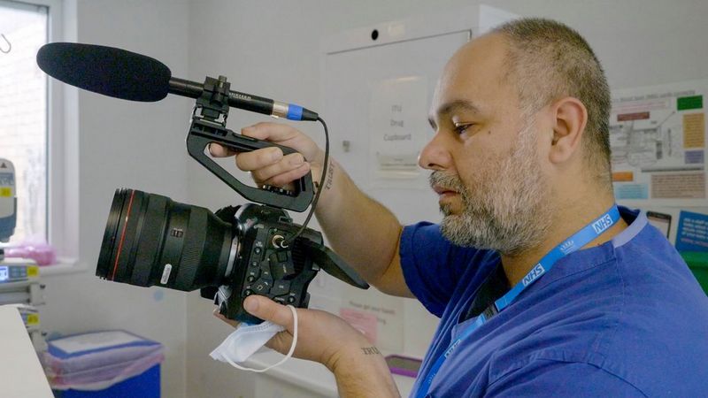 Giulio Di Sturco wearing hospital scrubs and holding up a Canon EOS C70 with an external microphone attached.