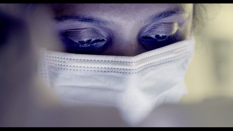 A still from a video shot on a Canon EOS C70, showing a close-up of the face of a healthcare worker wearing a surgical mask covering her mouth and nose.