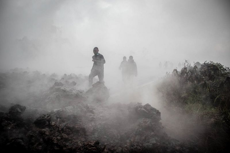 A group of people emerging from thick white smoke, walking across black jagged rocks and rubble. 