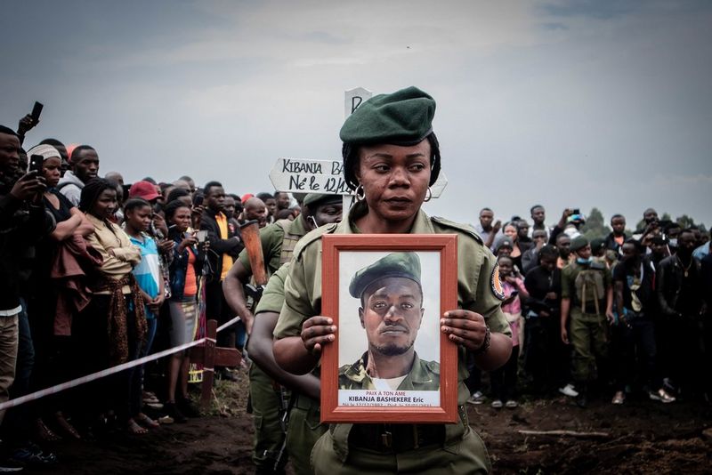 A funeral procession: a woman in uniform with a green beret holds a portrait of a man in the same uniform while bystanders stand solemnly, many taking photographs. 