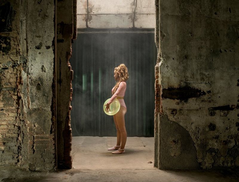 Incorniciata da una porta fatiscente, una modella con una maschera barbuta in silicone tiene in mano un palloncino giallo trasparente e guarda desolata verso il basso.