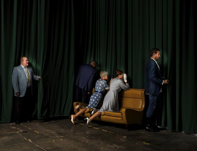 Un gruppo di uomini e donne in abiti eleganti stile anni '50 sbircia attraverso un sipario verde.