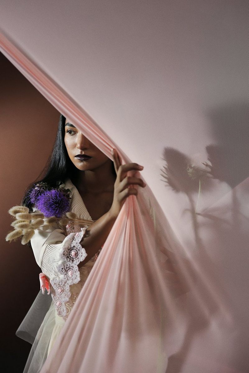 Una giovane donna dai capelli neri regge un piccolo mazzo di fiori e sbircia da dietro un sottile telo rosa.