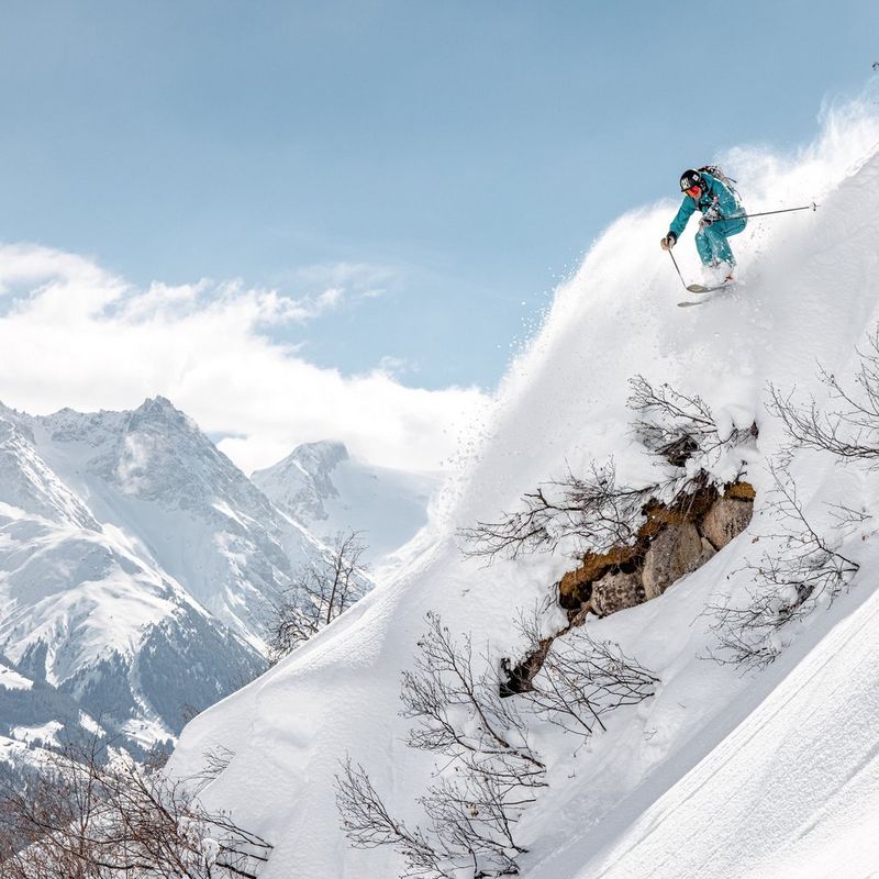 A skier dressed in turquoise on the edge of a steep slope, with snow-covered mountains in the background. 