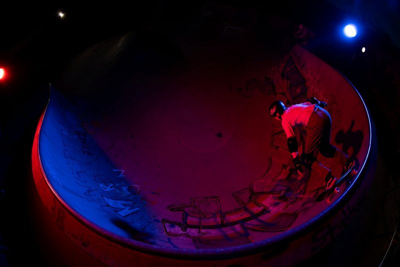 Een foto, genomen met een fisheye-objectief, van een meisje dat 's avonds skateboardt op een schans met rode en paarse verlichting.
