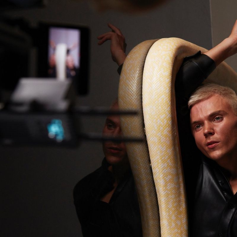 A Canon Cinema EOS camera films a young man with short blond hair lying on the floor. An enormous, yellow Burmese python is coiled around his raised arm