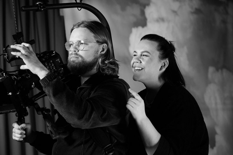 A black and white image of a man adjusting the display screen on a Canon Cinema EOS camera, while a woman looks over his shoulder smiling.
