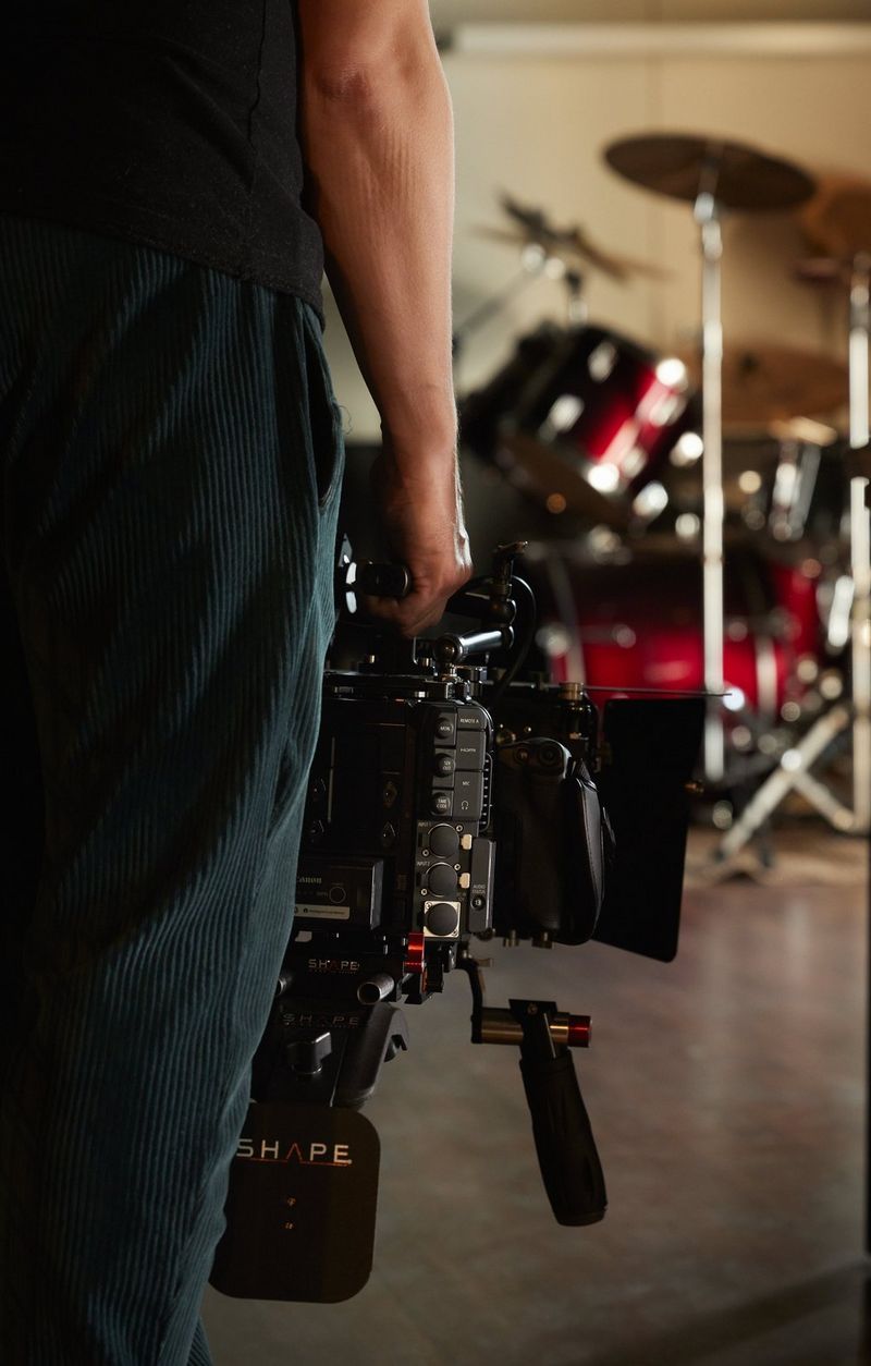 A person holding a Canon Cinema EOS camera. In the background is an elaborate drum kit.