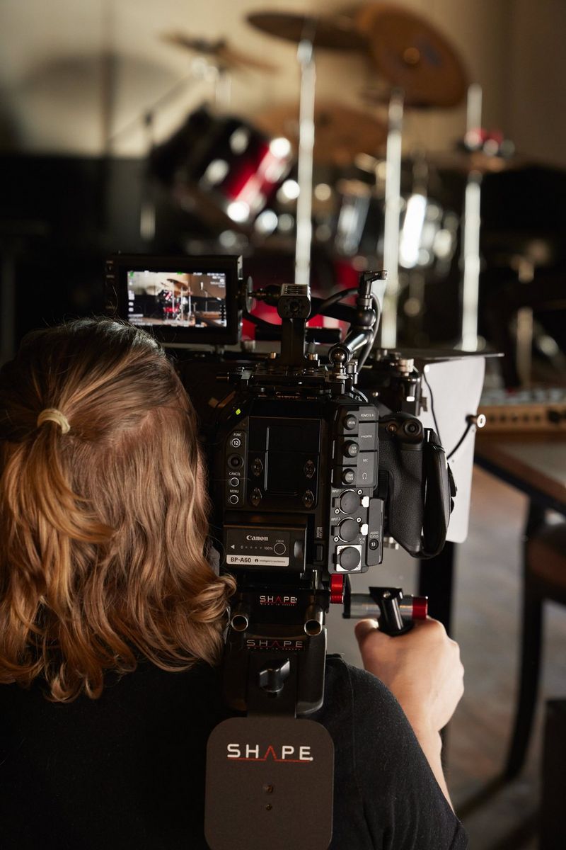 A long-haired man, with his back turned, films a drum kit with a Canon Cinema EOS camera.
