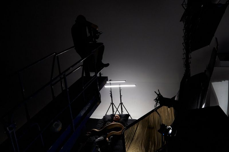 A young man with a large Burmese python coiled on top of him is filmed from above on a music video set. Aside from two thin strips of light, the studio is in darkness.