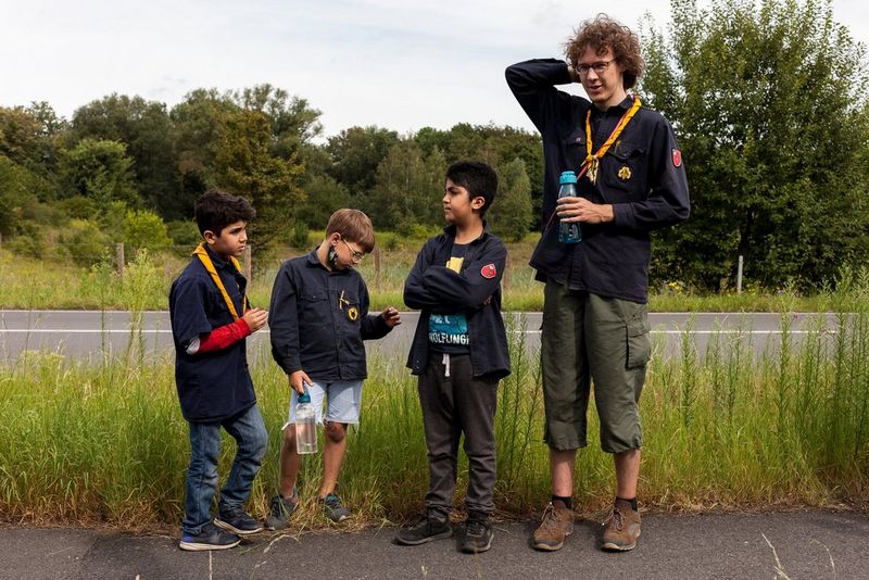 Drie jonge scouts en een oudere scoutingleider staan te wachten langs de kant van een landweg.