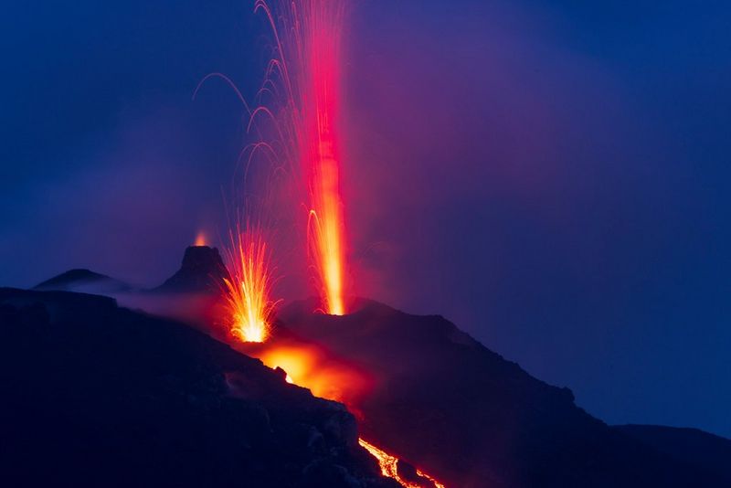 Черные силуэты вулканов на темно-синем фоне с яркими красными и оранжевыми искрами и струями лавы.