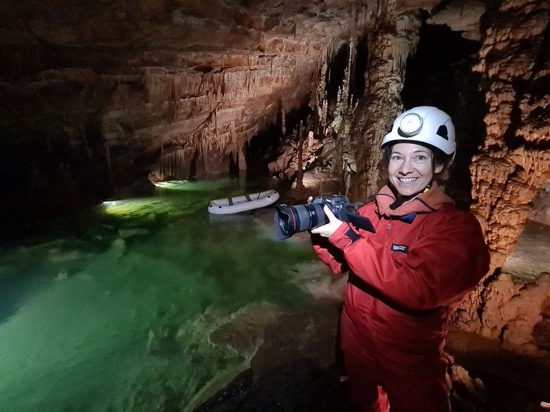 Улла позирует с камерой в подводной пещере, на ней надет красный водонепроницаемый костюм, белая каска и фонарь.