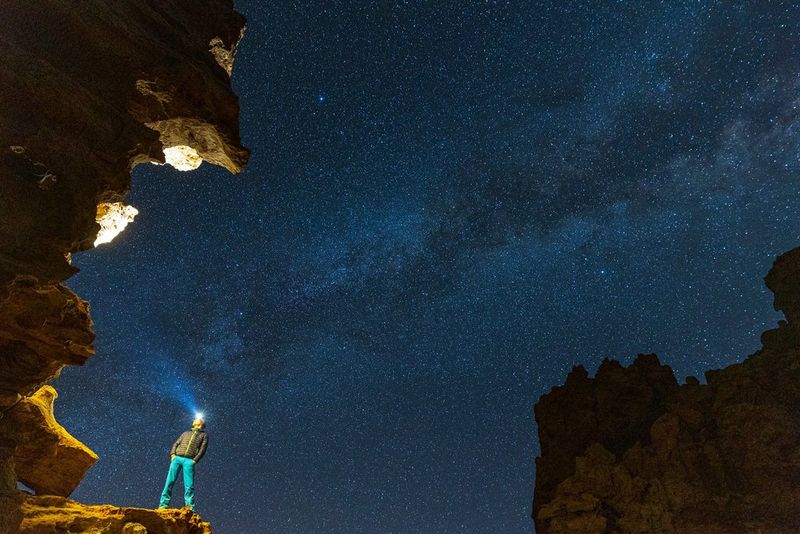 Мужчина с налобным фонарем стоит на скальном козырьке, сложив руки в карманы. Большую часть изображения занимает ночное небо, на котором видны тысячи звезд.