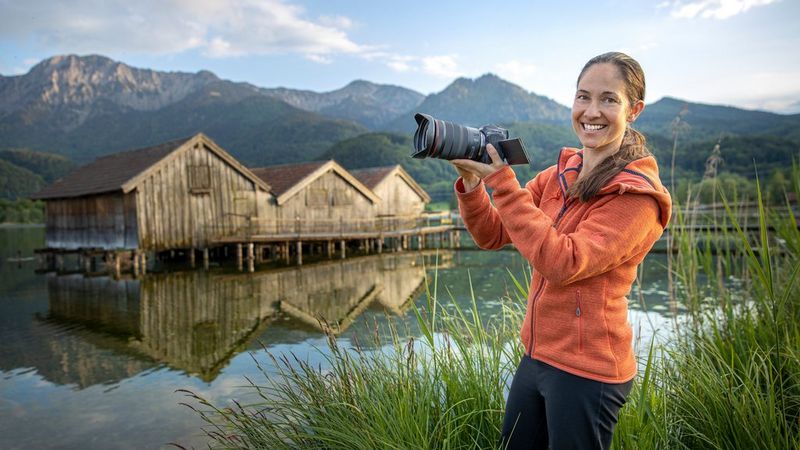 Фотограф приключений и путешествий Улла Лохманн стоит с камерой Canon EOS R5 перед деревянной хижиной на сваях в реке.