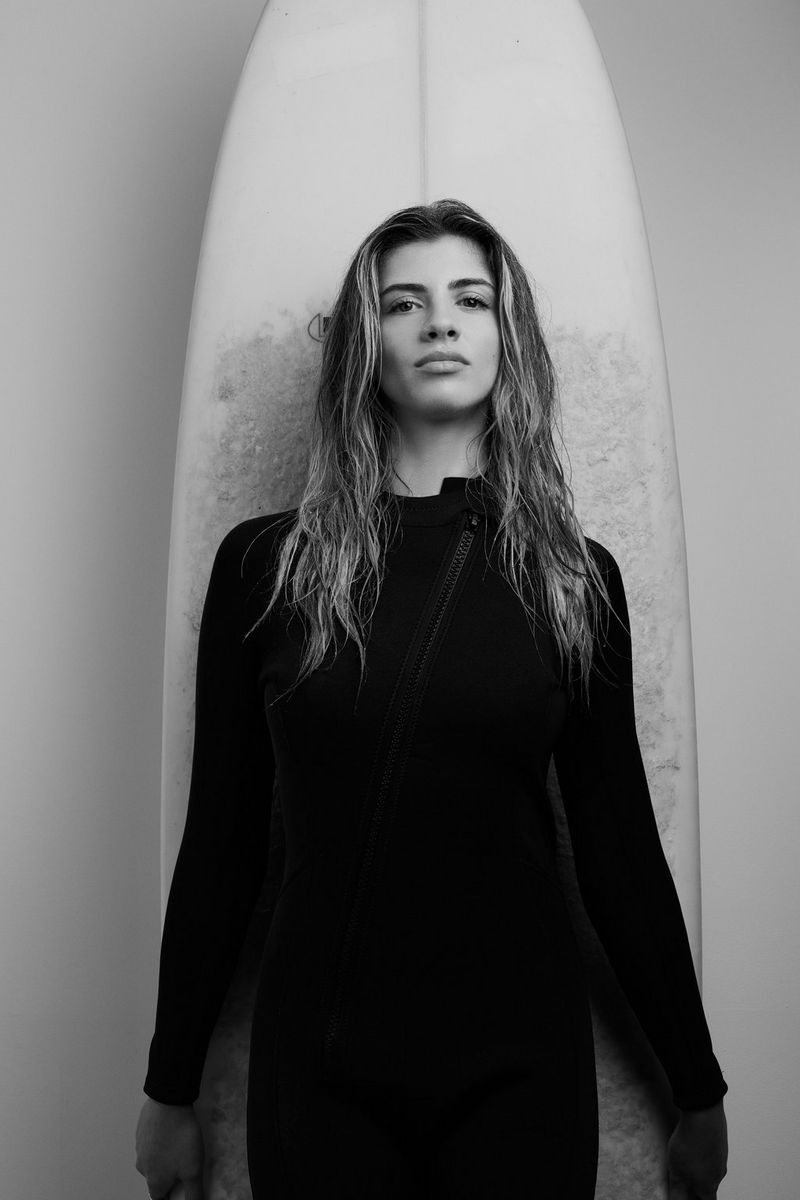  A model wearing black clothing stands against a surfboard in a studio, with the portrait converted to monochrome. Taken using a Canon RF 85mm F1.4L VCM hybrid lens