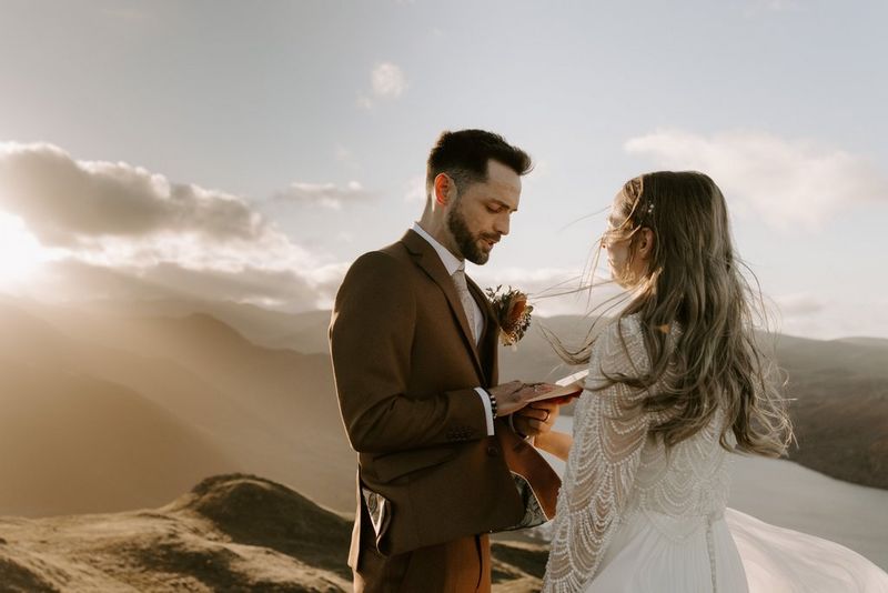 A formally dressed bride and groom face each other on a hilltop, with a rugged landscape in the background, lit by the sun’s rays through a cloud on one side. Taken on a Canon EOS R5 by wedding photographer and videographer Jo Greenfield.