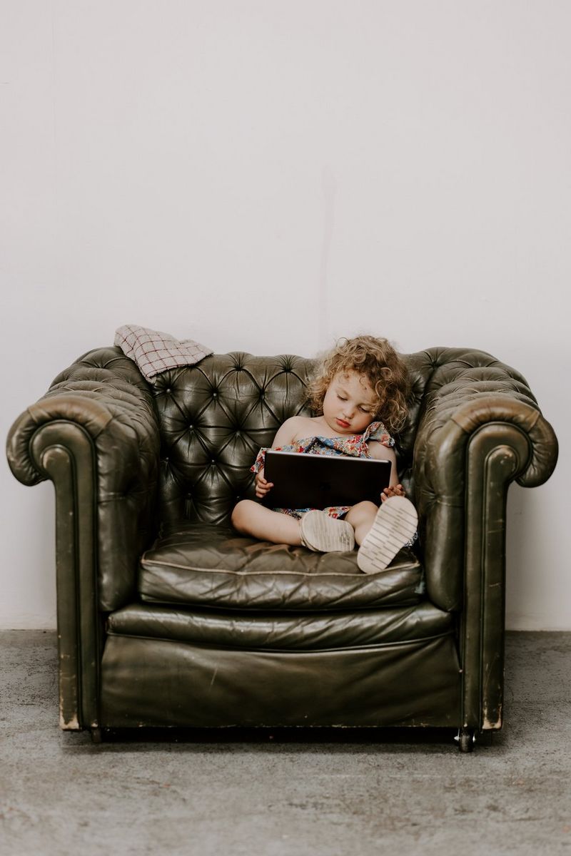 A little girl dozes in a large green upholsteed armchair, a tablet held loosely in her fingers. Taken on a Canon EOS R5 by wedding photographer and videographer Jo Greenfield.