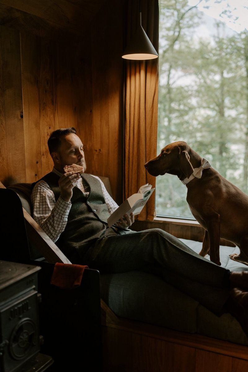 A man in a waistcoat relaxes in a window seat reading a book, with a large brown dog sitting alongside him, staring intently at the slice of bread he is holding in his othe hand. Taken on a Canon EOS R5 by wedding photographer and videographer Jo Greenfield.