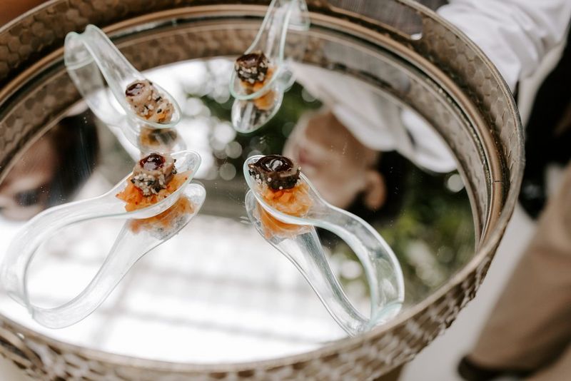 Four elaborate hors d’ouevres on curved transparent spoons, sitting on a silver serving tray, with the waiter’s head reflected in the surface. Taken on a Canon EOS R5 by wedding photographer and videographer Jo Greenfield.