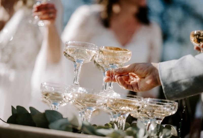A hand taking a glass of champagne from a stack, with a woman in a white dress visible in the background. Taken on a Canon EOS R5 by wedding photographer and videographer Jo Greenfield.