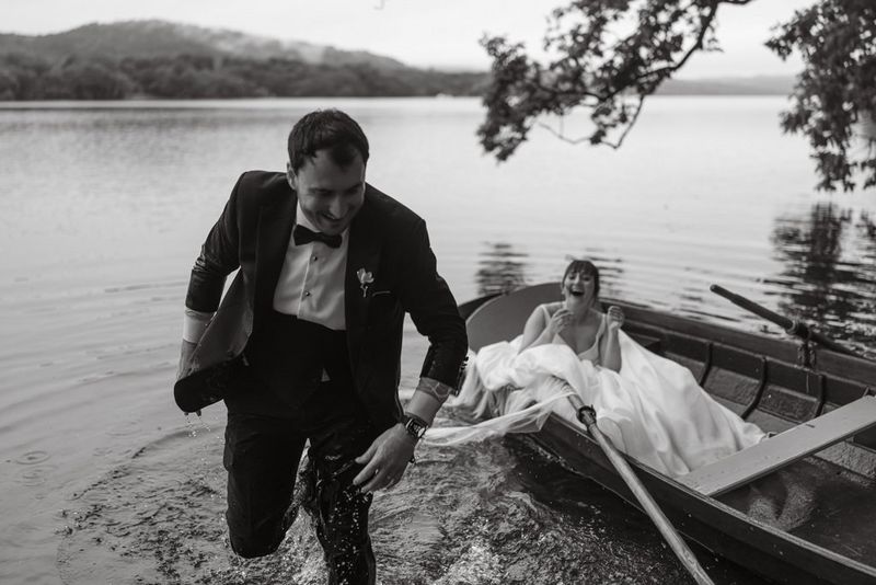 A formally dressed groom, soaking wet, stands in a lake, with the bride in a rowboat behind him laughing with him. Taken on a Canon EOS R5 by wedding photographer and videographer Jo Greenfield.