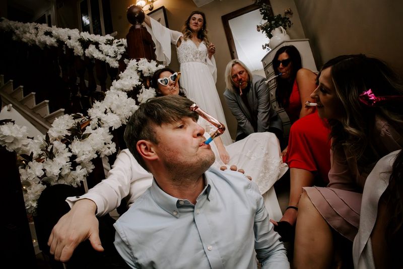 Guests blow party horns in a lively wedding celebration on a sweeping staircase adorned with white flowers all the way up the handrail. Taken on a Canon EOS R5 by wedding photographer and videographer Jo Greenfield.