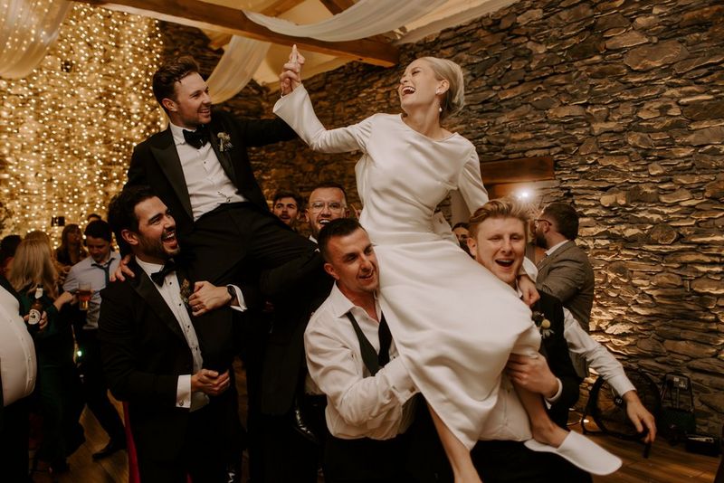 A laughing bride and groom are carried on the shoulders of guests at the wedding reception. Taken on a Canon EOS R5 by wedding photographer and videographer Jo Greenfield.
