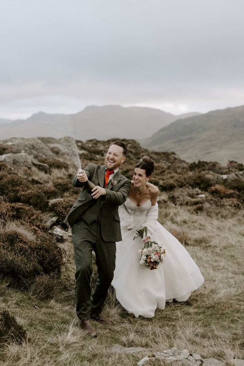 A formally dressed groom in a rugged Highland landscape sprays champagne from a bottle, while the bride, wearing a white wedding gown and holding a bouquet, laughs with him. Taken on a Canon EOS R5 by wedding photographer and videographer Jo Greenfield.