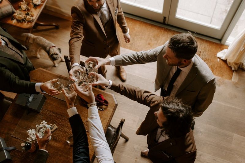 A top-down view of a groom and male wedding guests clinking their glasses together in a toast. Taken on a Canon EOS R5 by wedding photographer and videographer Jo Greenfield.