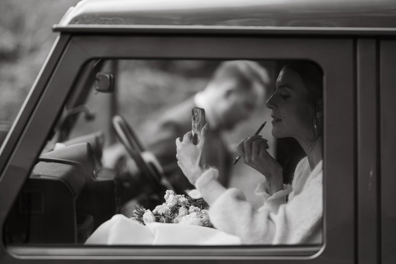 Black and white photo of a bride sitting in the passenger seat of a Land Rover, touching up her makeup. A male figure is out of focus outside the car on the other side. Taken on a Canon EOS R5 by wedding photographer and videographer Jo Greenfield.