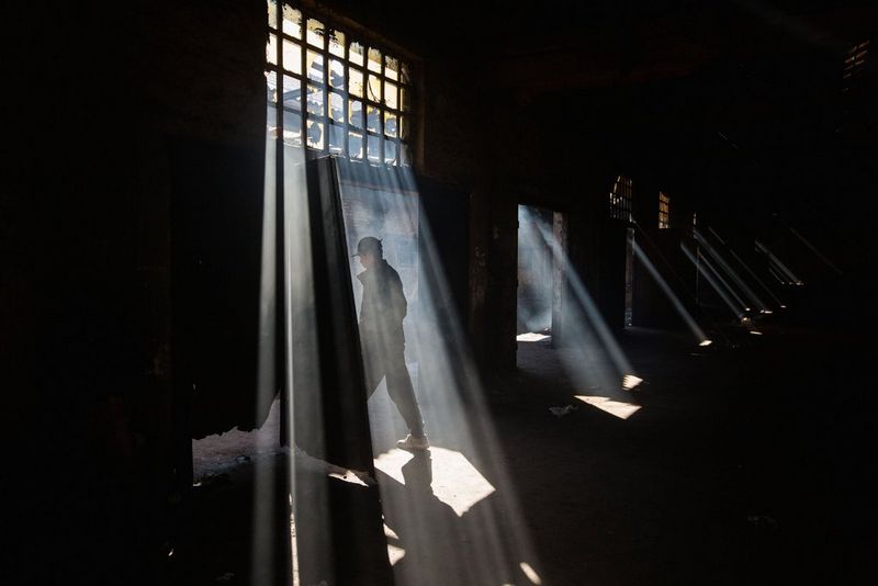 A man exits a dark warehouse as sunlight streams through the broken windows. Photo by Jędrzej Nowicki