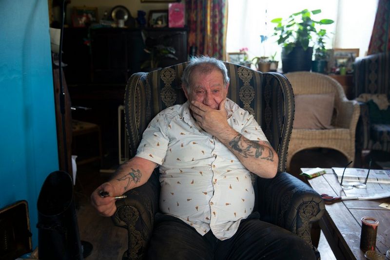 Мужчина сидит в кресле, приложив одну руку ко рту, а в другой удерживая трубку; на его руках можно увидеть татуировки.