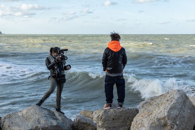 Filmmaker Francesca Tosarelli uses a Canon EOS C70 camera to film her subject, with both standing on rocks on the seashore.