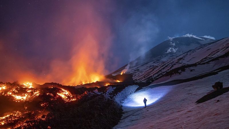A view of a lone climber standing on a snowy mountainside filming lava flowing during an eruption at Mount Etna, Sicily. 