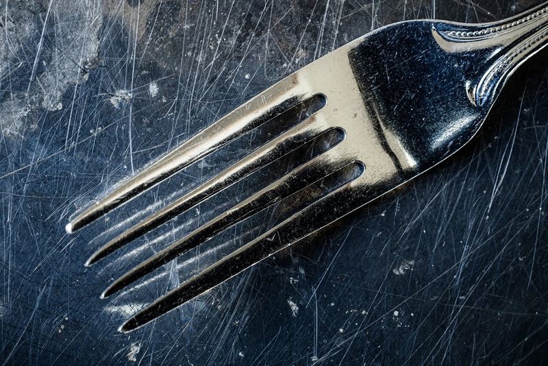 A table fork photographed on a dark work surface with numerous scratches visible in it.