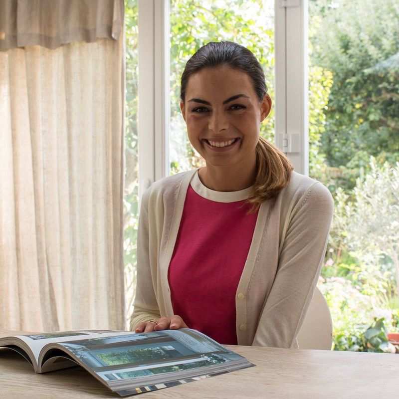 A young woman sits at a table with a window behind her, so that her face is in shadow.