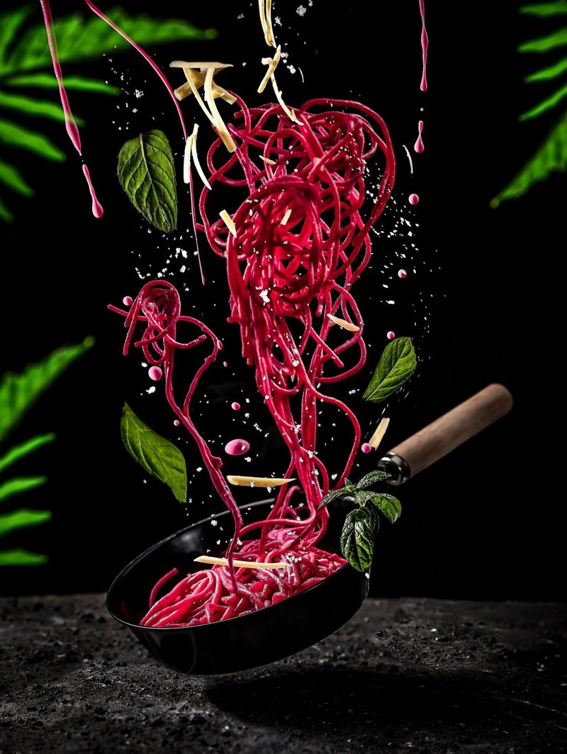 Pink spaghetti falling into a pan suspended above a counter, in front of a black background and framed by bright green foliage.