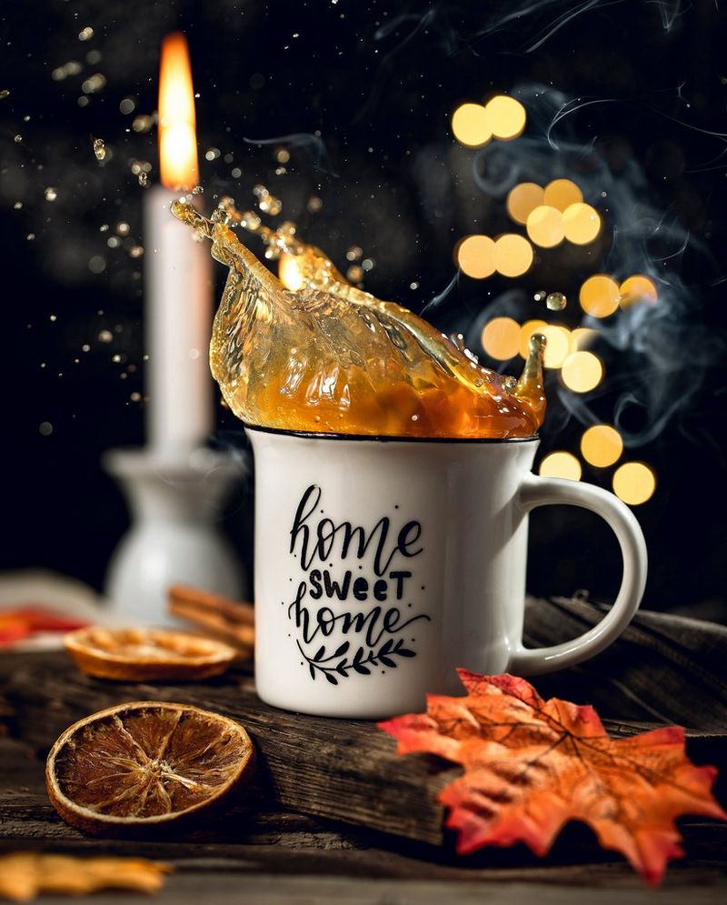 Liquid splashes upwards from a mug that sits on a dark wood board surrounded by dried fruit and autumn leaves. A candle is burning in the background.