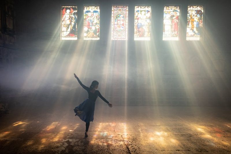 A woman dancing lit by beams of sunlight falling through a row of stained glass windows.