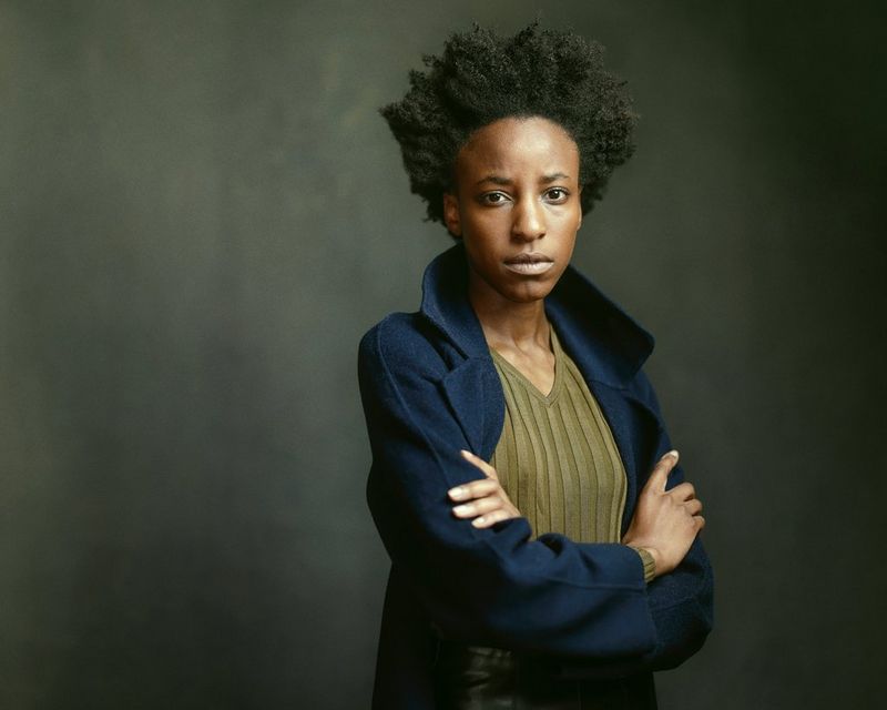 Una ragazza dai folti capelli scuri con un cappotto in lana a collo alto fissa direttamente la fotocamera, con le braccia incrociate sul petto.